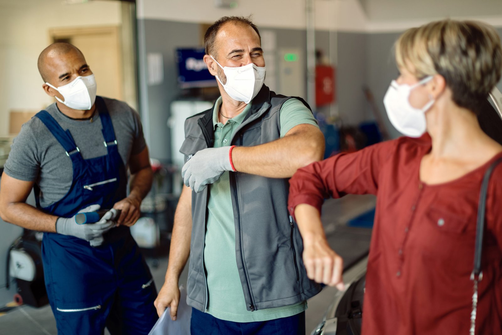 Auto repairman and his customer wearing protective face masks and greeting with elbows in a workshop.